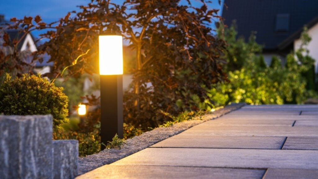 Soft, warm light from a modern bollard lamp brightens a garden walkway and nearby shrubs as evening sets in.