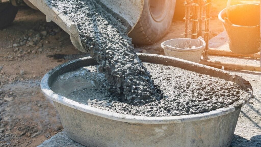 A close-up shot of wet, ready-mix concrete being discharged from the chute of a concrete truck into a large, grey plastic or metal mixing tub on a construction site.