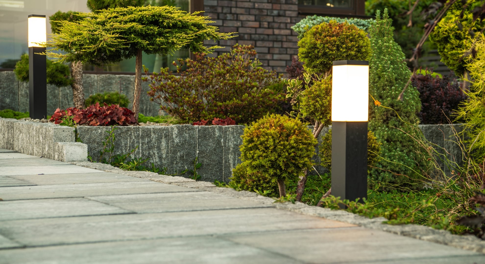 A modern outdoor space with a stone pathway, illuminated by a tall lamp post, surrounded by shrubs and trees