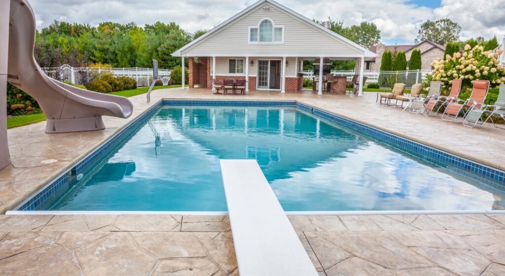 A rectangular swimming pool with a diving board, surrounded by a patio and a large house with a covered outdoor area