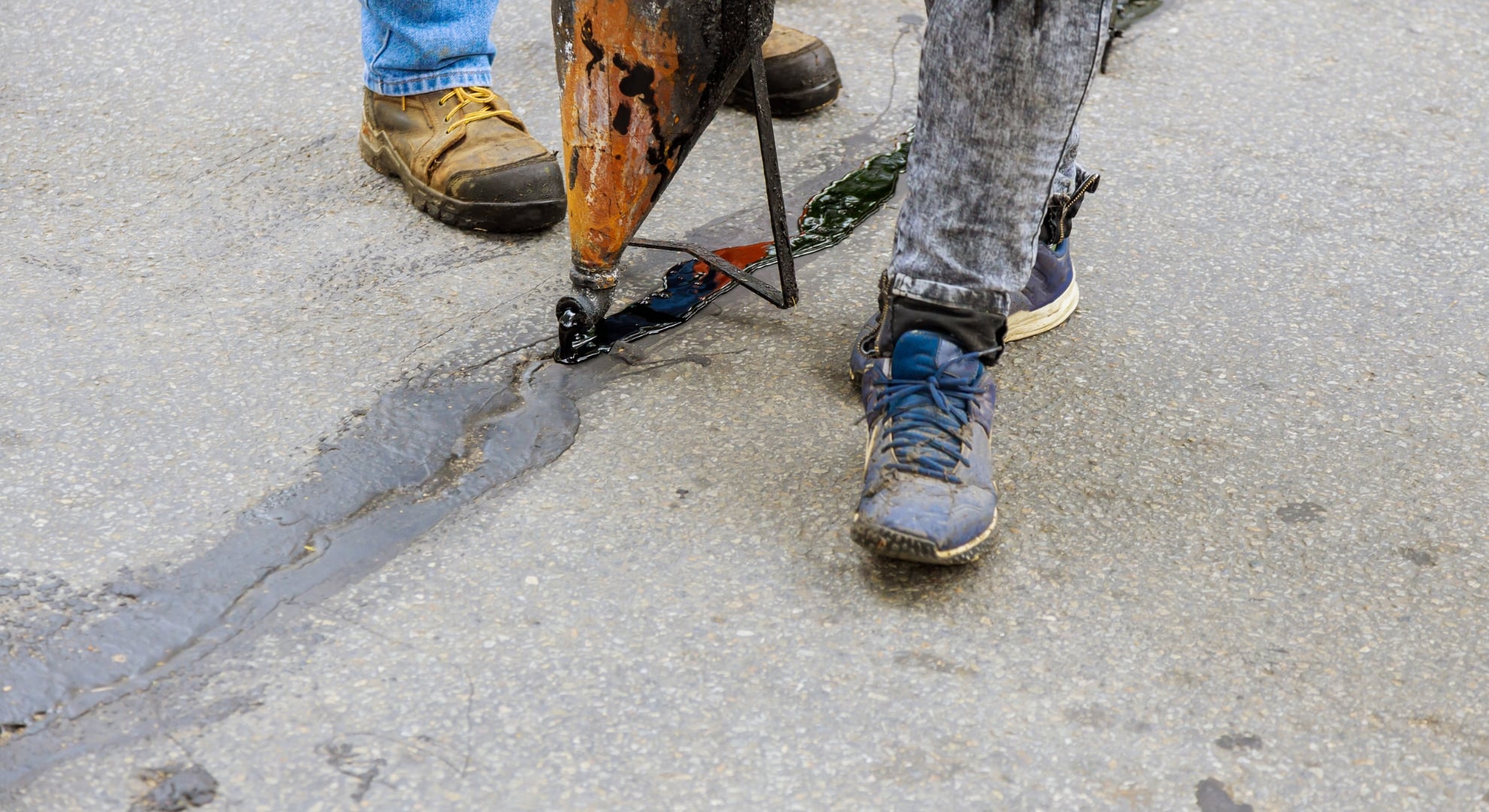 Two workers filling a crack in concrete with a hot tar sealant.