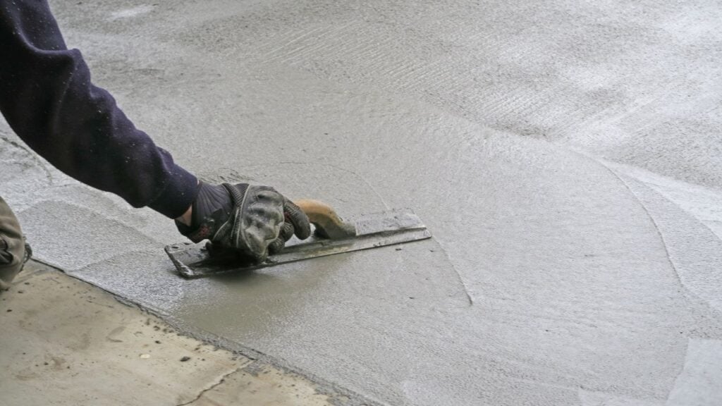 A close-up view of a worker, wearing dark gloves, using a flat metal trowel to smooth and apply a concrete patch or fresh concrete onto an existing surface.