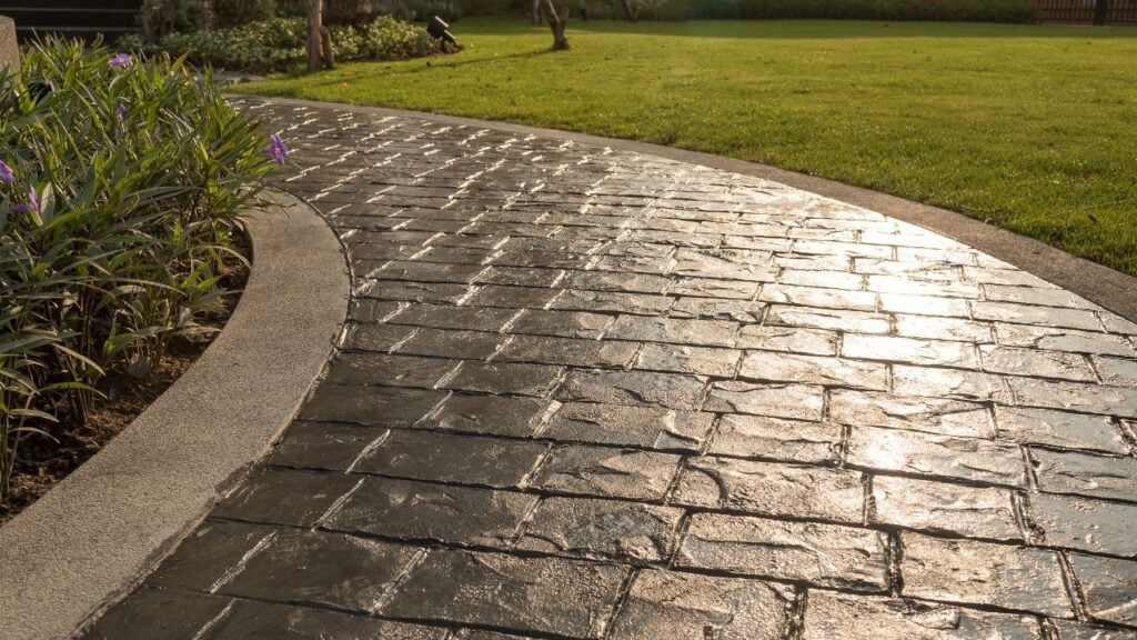 A curving outdoor concrete walkway featuring a dark grey, glossy stamped design that mimics a herringbone brick pattern.