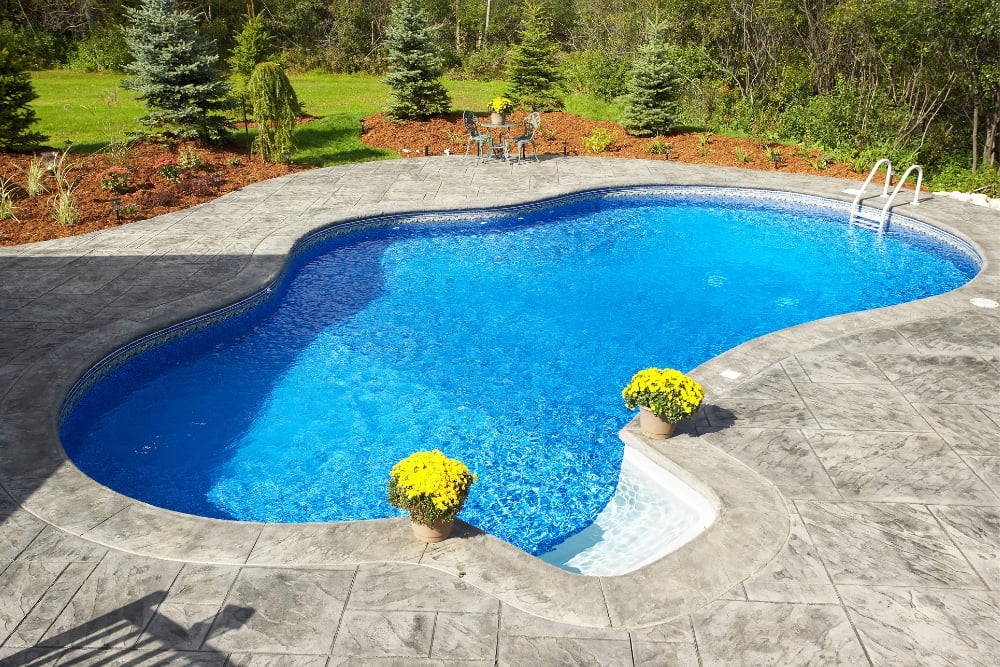 A bright blue inground swimming pool surrounded by grey honed concrete paving in a landscaped backyard.