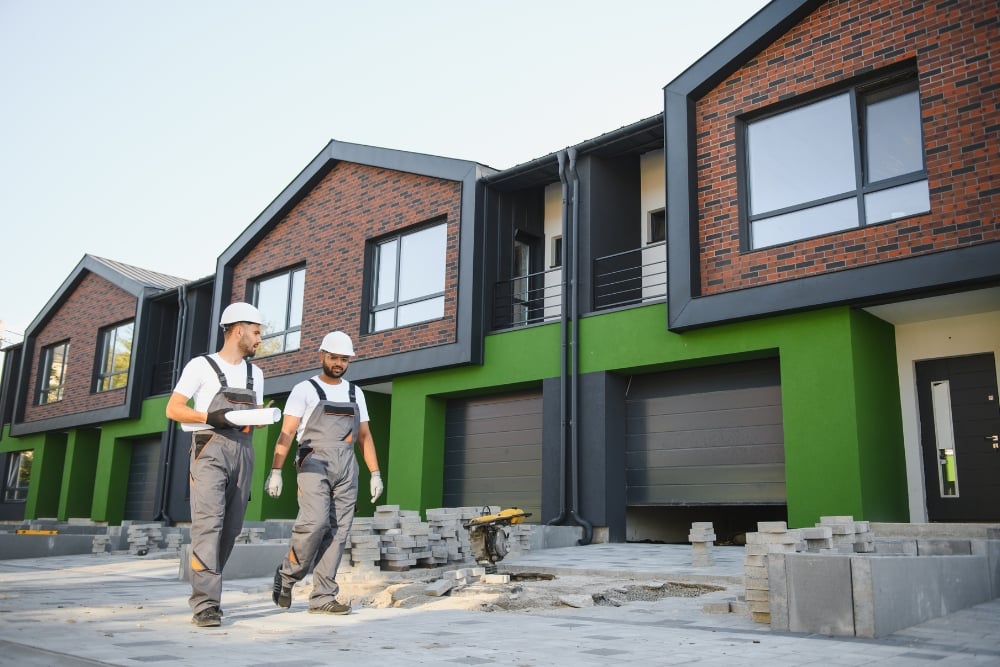 Two builders in hard hats and overalls walking on a paved driveway outside new residential housing units.