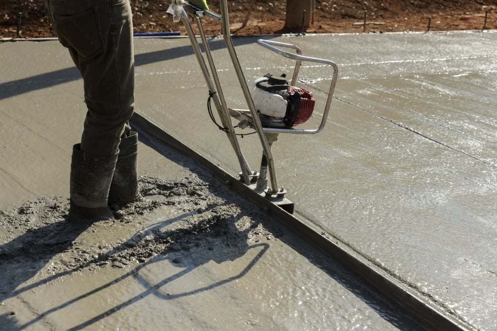 A contractor in rubber boots using a power screed machine to smooth and level fresh wet concrete.