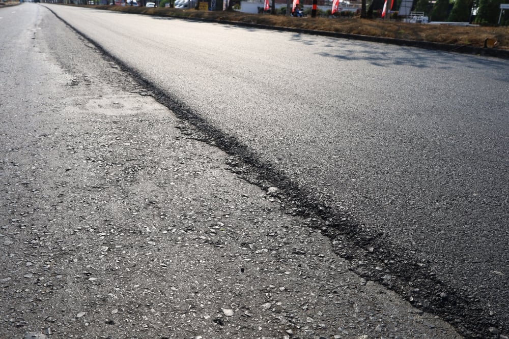 An asphalt road showing a distinct dividing line where a fresh, smooth layer of dark asphalt meets an older, rougher, and worn-down road surface.