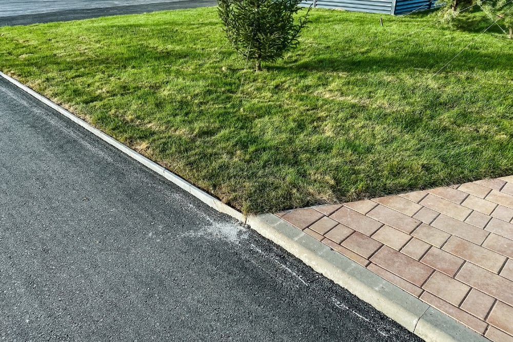 A newly paved asphalt surface bordering a stone curb, a green lawn, and a section of interlocking brick pavers.