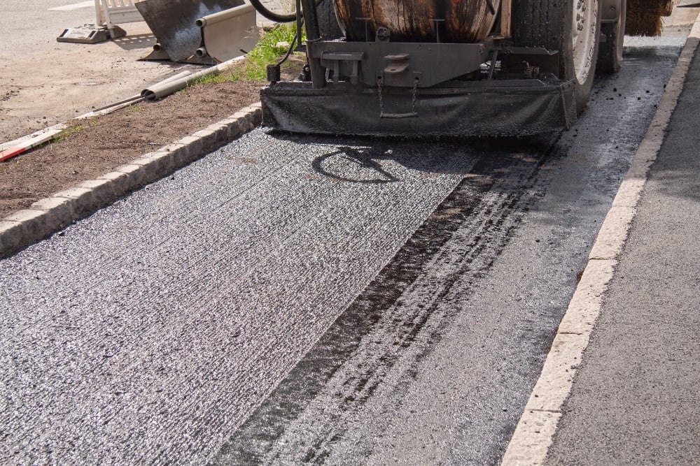 A paving machine resurfacing a fresh, dark layer of textured asphalt onto a narrow driveway flanked by stone curbs.