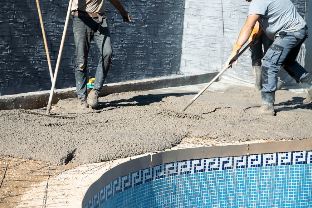 Construction workers using rakes to spread and level wet concrete over a wire mesh grid along the edge of a blue-tiled swimming pool.