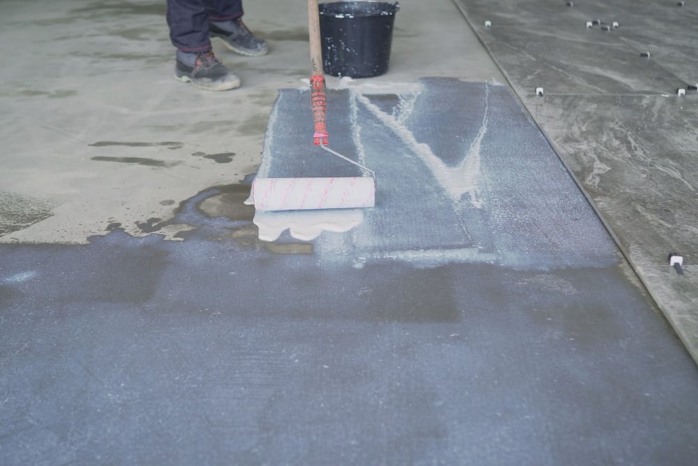 A worker uses a paint roller to apply a white liquid primer or adhesive to a grey concrete slab.