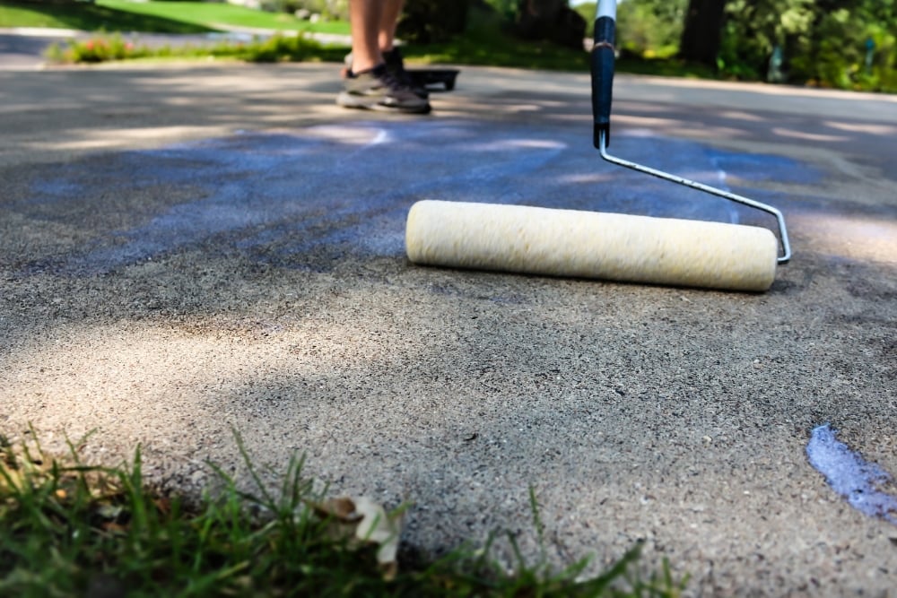 A paint roller applying a blue-tinted sealant coating onto a textured concrete driveway surface, with a worker's feet visible in the blurred background.
