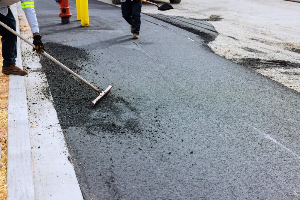 A construction worker uses a long asphalt lute rake to smooth down fresh asphalt patch mix along a concrete curb.