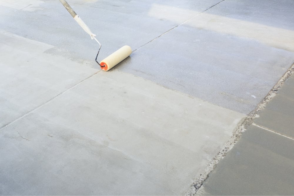 A paint roller applies a clear, glossy liquid sealer to a smooth gray concrete slab divided by expansion joints.