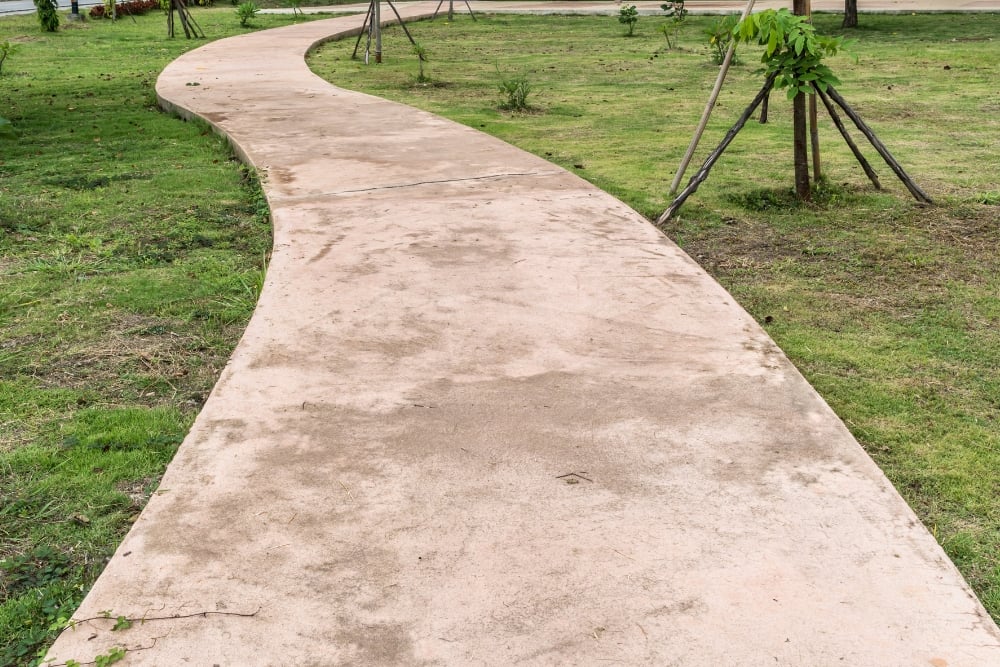 A winding, light-pink tinted concrete pathway curving through a green grassy park area.