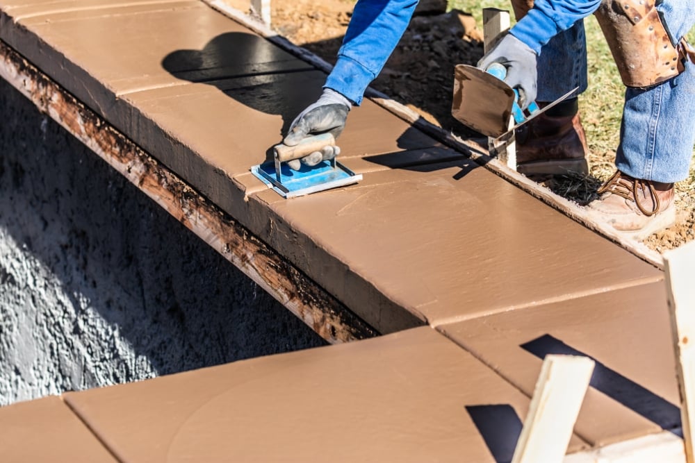 A construction worker wearing gloves uses hand trowels to smooth and shape wet, brown concrete along the top edge of a pool bond beam.