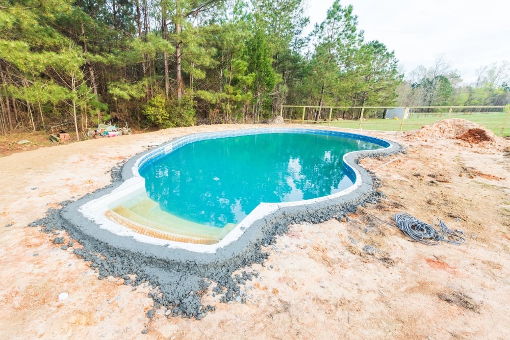 An inground swimming pool filled with turquoise water under construction, featuring a freshly poured gray concrete bond beam lining its curved edge in a dirt yard.