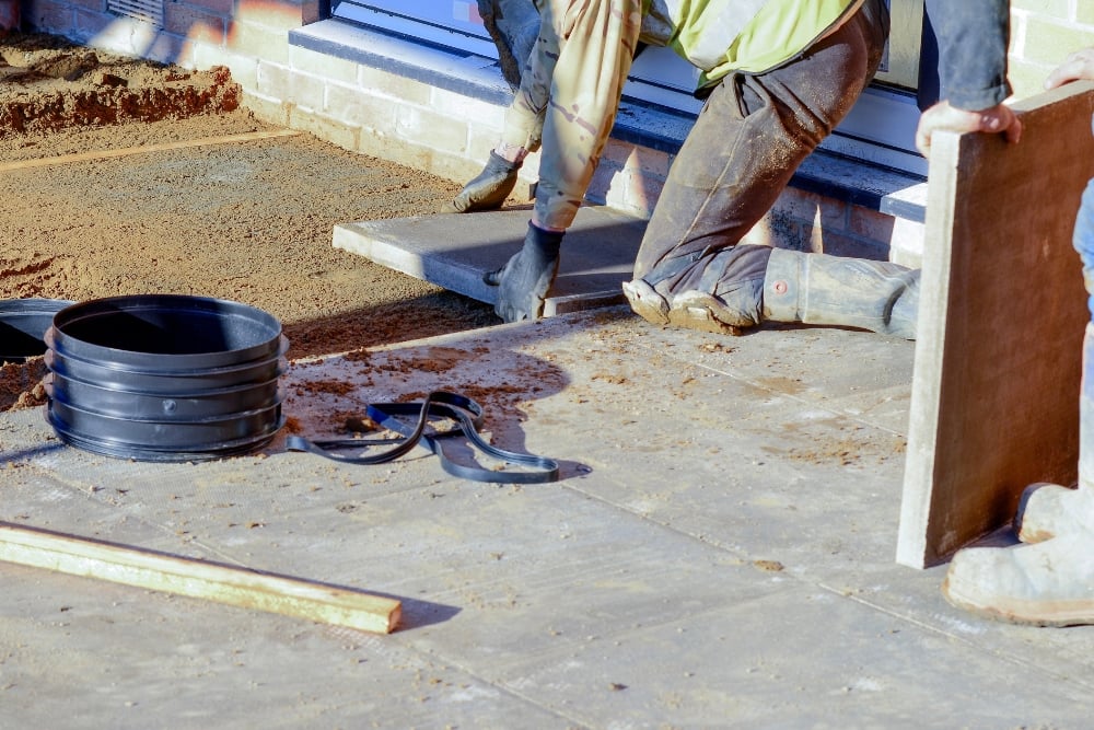 Construction workers on a dirt site kneeling down to position rigid insulation boards over a ground layer next to a building foundation.