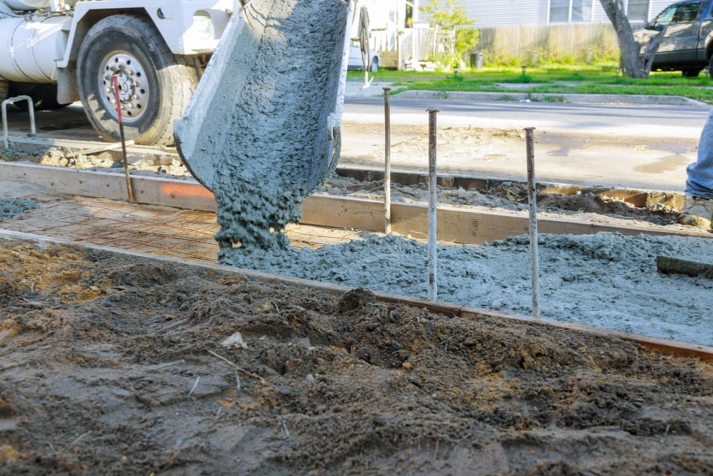 A ground-level view of wet concrete pouring from the metal chute of a mixing truck into a wooden form lined with steel mesh rebar for a new sidewalk or driveway construction.