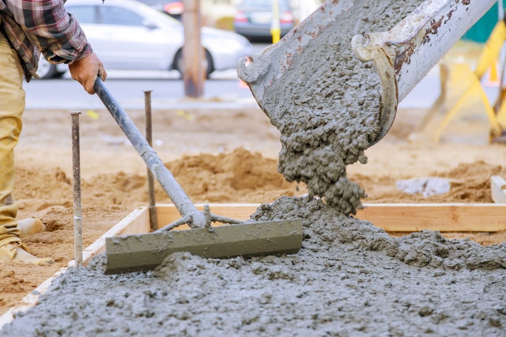 Construction worker spreading wet concrete poured from a mixer chute using a rake.