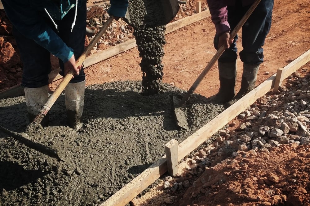 Two workers spreading fresh wet concrete into wooden formwork with hand tools.