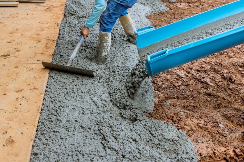 A construction worker wearing rubber boots uses a rake to spread fresh, wet concrete pouring from a blue chute.