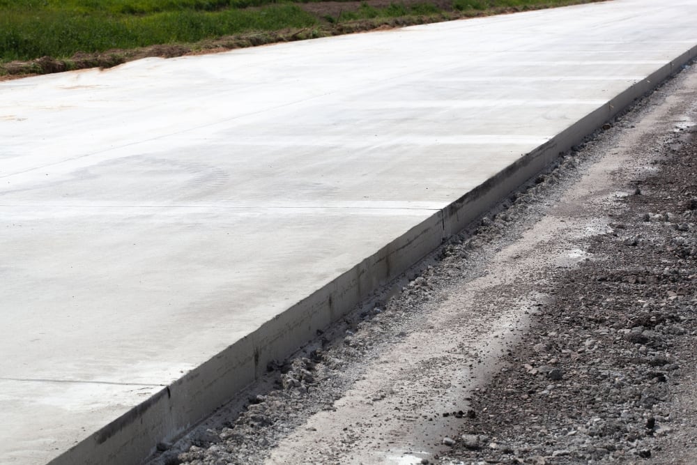 A newly laid, smooth concrete pavement with a clean vertical edge running alongside a dirt and gravel shoulder.