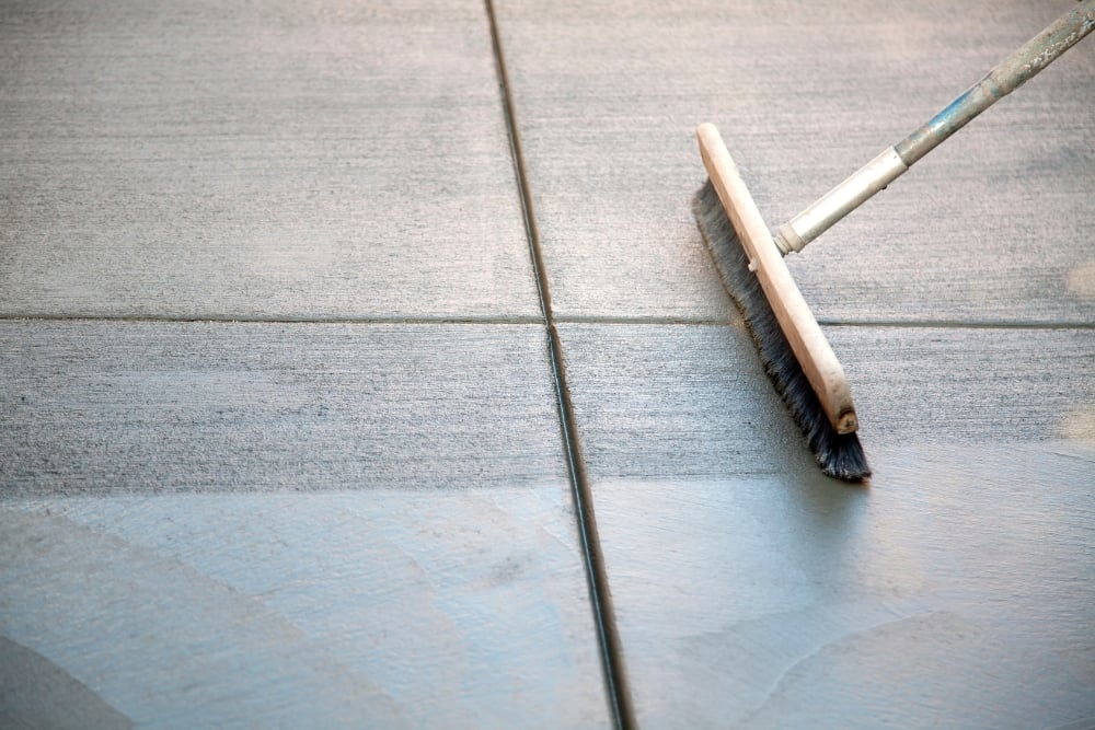A close-up view of a broom being used to create a textured finish on freshly laid or wet grey concrete slab panels.