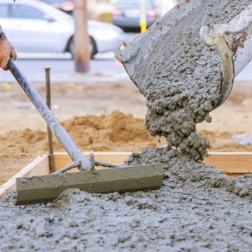 Construction worker spreading wet concrete poured from a mixer chute using a rake.