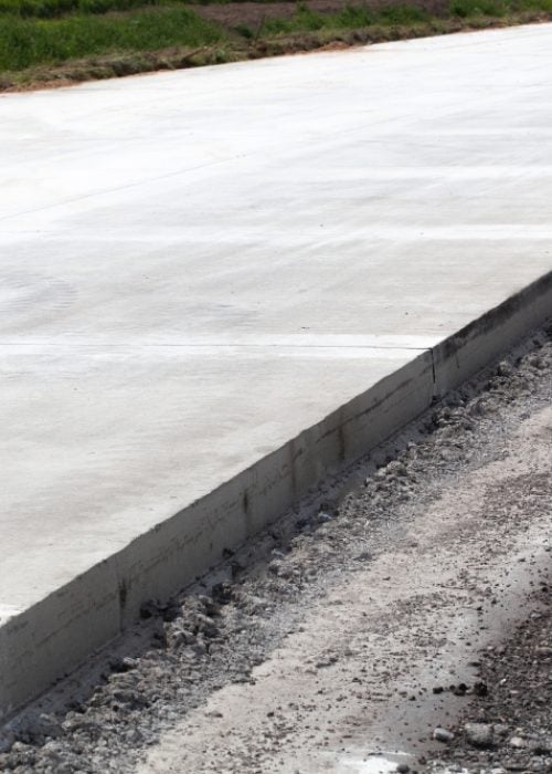A newly laid, smooth concrete pavement with a clean vertical edge running alongside a dirt and gravel shoulder.